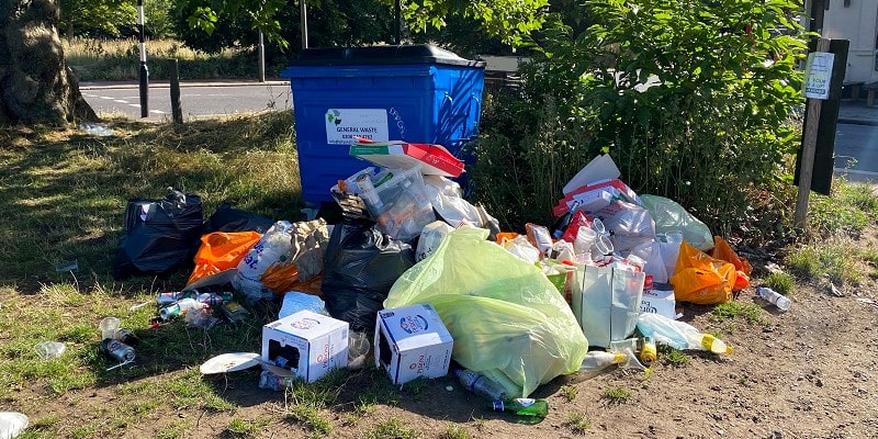 Large amount of household rubbish illegally dumped around a blue commercial waste container.