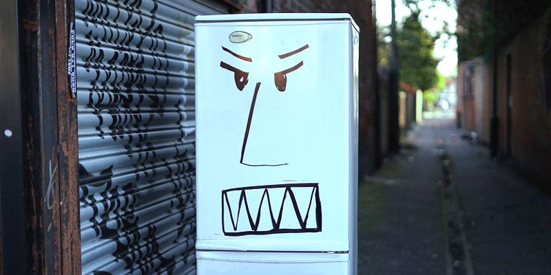 White fridge placed in the middle of a city service lane. Fridge has an angry face drawn on it.