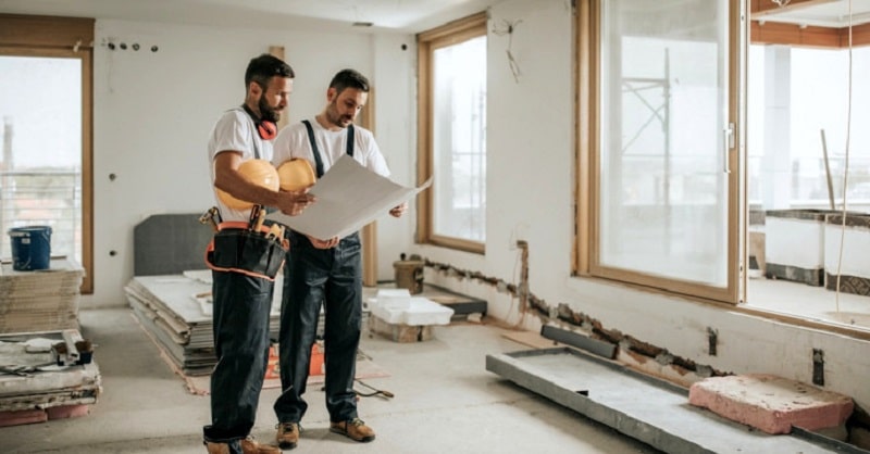 Two male builders looking at building plans inside a partially renovated room